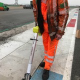 Werknemer in oranje werkkleding voert een meting uit langs de rand van een asfaltweg.